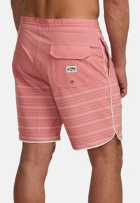 Ružové plavky s pruhovaným vzorom, elastickým pásom, bočnými a zadnými vreckami, a nášivkou loga na zadnom vrecku.