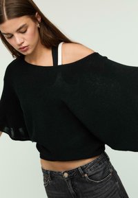 Pull en tricot noir oversized avec un décolleté bateau, doté de manches larges et d'une finition texturée, porté par-dessus un débardeur noir et blanc.