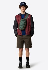 Giacca leggera color bordeaux e nera sopra una camicia blu, pantaloni corti oliva e una borsa a tracolla verde. Include calze scure e scarpe nere.