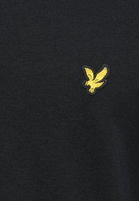 Camiseta de algodón negra con un pequeño logo de pájaro bordado en amarillo en la parte izquierda del pecho. Textura suave, escote redondo clásico.
