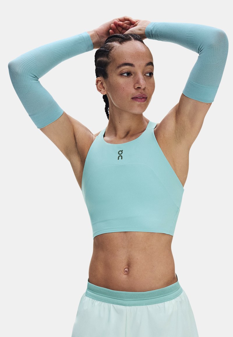 Personne athlétique portant un haut de sport bleu clair et des manchons, étirant les bras au-dessus de la tête, regardant sur le côté.