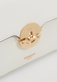 Valkoinen nahkainen käsilaukku, jossa on kultainen pyöreä solki ja kohokuvioitu logo, sekä sileä pinta ja kontrastina mustat ompeleet.