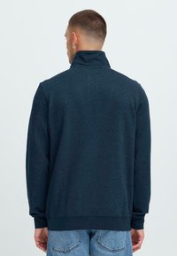 Moški s kratkimi lasmi in uhani, oblečen v temno modro pulover z visokim ovratnikom in svetlo modre kavbojke, obrne se stran proti enobarvni ozadju.