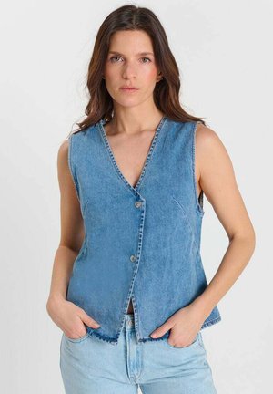 Vrouw met bruin haar draagt een mouwloos blauwe spijkervest en lichtblauwe jeans, staand met handen in de zakken tegen een effen achtergrond.