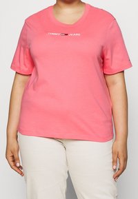 Camiseta de algodón rosa con mangas cortas, escote redondo y logo "TOMMY JEANS" en blanco y azul marino. Estilizada con pantalones de color crema.