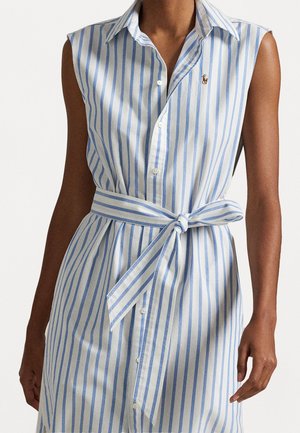 Frau trägt ein ärmelloses blau-weiß gestreiftes Hemdkleid mit vertikalen Streifen, einem gebundenen Gürtel in der Taille und Knöpfen vorne.