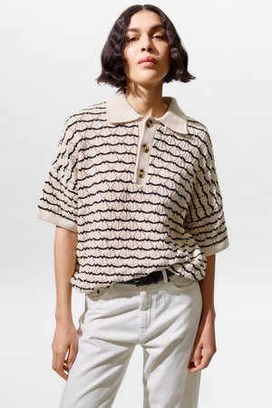 Jauna moteris su trumpais bangeliais plaukais dėvi smėlio spalvos ir juodos spalvos raštuotą megztą polo marškinėlį, įkištą į baltas kelnes su juodu diržu.