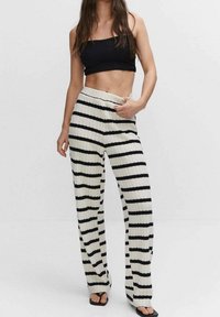 Femme portant un crop top noir et un pantalon en tricot blanc taille haute avec des rayures noires horizontales, se tenant devant un fond uni.