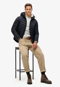 Chaqueta negra acolchada con capucha, sudadera gris, pantalones caqui y botas marrones. Modelo sentado en un taburete de metal negro. Fondo mínimo.