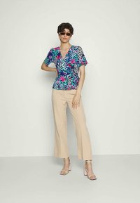Blusa floral de várias cores com decote em V profundo e mangas curtas, combinada com calças largas bege claras e sandálias de salto rosa.