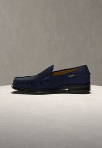 Μπλε ναυτικό σουέντ ανδρικό μοκασίνι με μαύρη σόλα, χαμηλό τακούνι, σχεδιασμό slip-on και διακοσμητικές ραφές σε ουδέτερο φόντο.