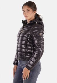 Giubbotto a ventosa nero con cappuccio, texture imbottita, chiusura con zip, dettagli in metallo dorato e un design aderente con una patch logo sulla manica.