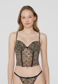 Žena v leopardím vzorovaném korzetu a odpovídajících tanga kalhotkách, stojící na jednofarebném světle šedém pozadí.