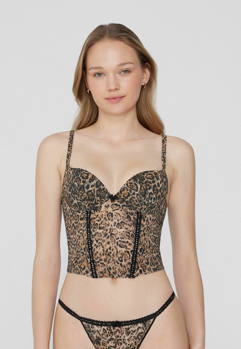 Žena v leopardím vzorovaném korzetu a odpovídajících tanga kalhotkách, stojící na jednofarebném světle šedém pozadí.