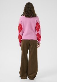 Różowy sweter wełniany z czerwonym wzorem argyle na rękawach, w połączeniu z brązowymi szerokimi spodniami z korduroy. Prosty, teksturowany i kolorowy design.