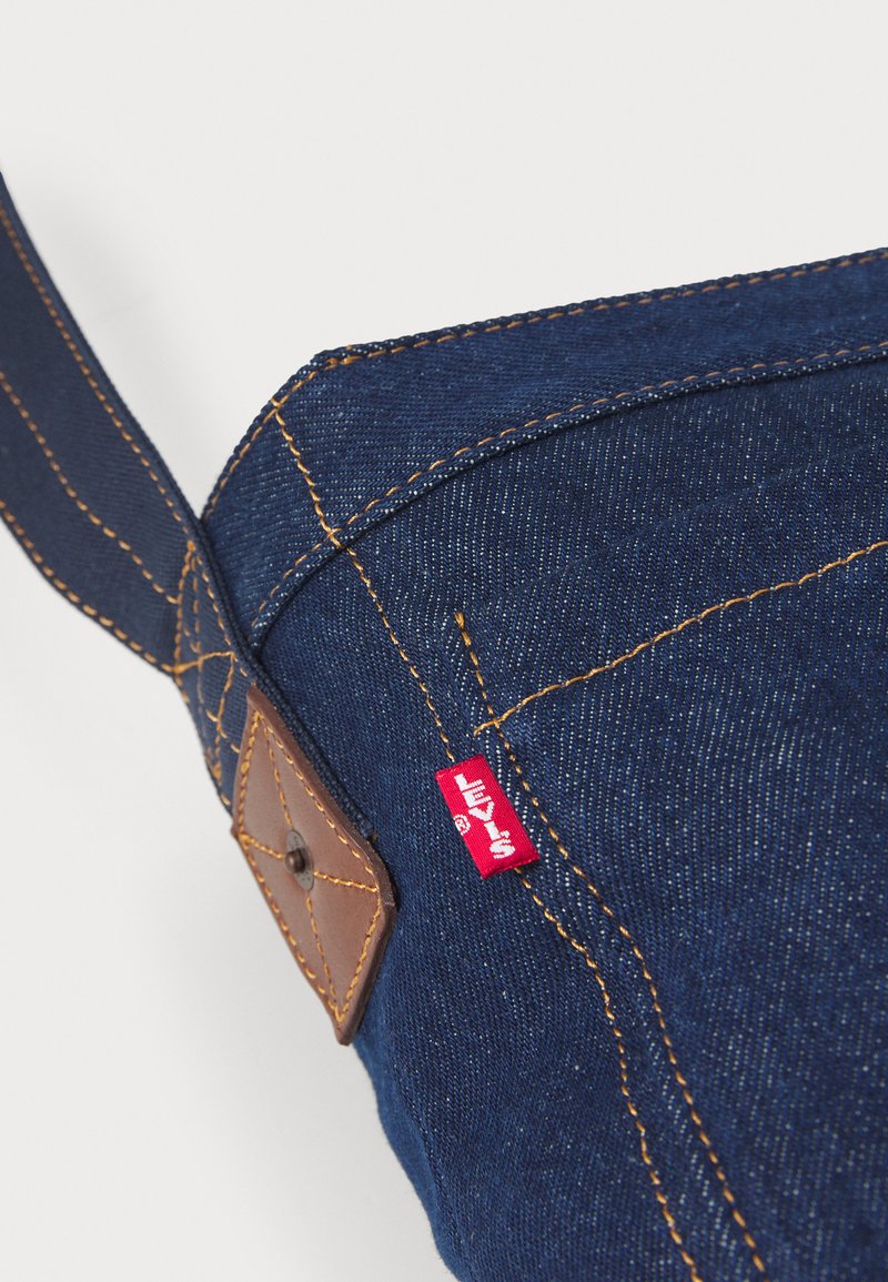 Denim stoffen tas in donkerblauw, met lichte stiksel en bruine leren accenten. Voorzien van een rode Levi's label en een gestructureerd ontwerp.