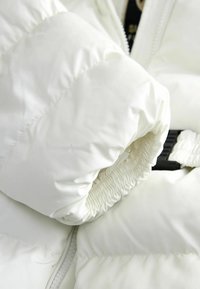 Balts pūkains džemperis tuvplānā, ar savilkta aproce, gludu tekstūru un redzamu rāvējslēdzēja detaļu ar melnām apdari.