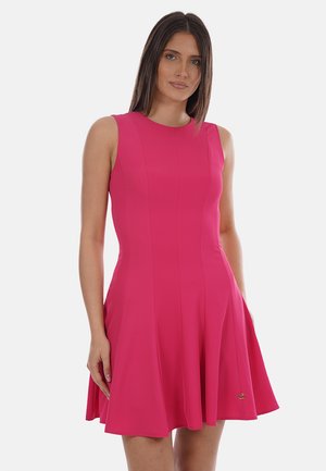 Donna che indossa un vestito aderente senza maniche, di colore rosa acceso, lungo fino al ginocchio con gonna a campana, in piedi contro uno sfondo chiaro e uniforme.