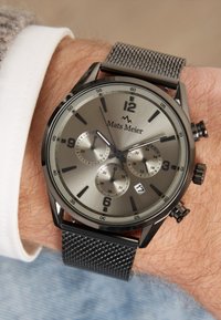 Montre bracelet en métal avec un cadran gris, trois sous-cadrans, un bracelet en maille noire et de grands index horaires. Comprend un logo et un affichage de la date.