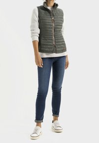 Olijfgroen, gewatteerde ritsen vest met een hoge kraag, gecombineerd met een crème, geribbeld sweater en blauwe jeans. Witte sneakers met zwarte accenten.