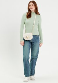 Mintgrüner gestrickter Pullover mit hohem Kragen, kombiniert mit blauen Jeans; das Model hält eine kleine weiße Umhängetasche mit runder Hardware.