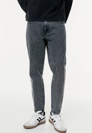 Personne portant un jean noir délavé et des baskets blanches avec des rayures noires, debout devant un fond uni.