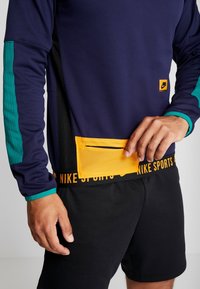 Maglia sportiva a maniche lunghe blu navy con maniche in accentuazione teal e tasca arancione in mesh. Pantaloni corti neri con vita stampata "NIKE SPORTS".