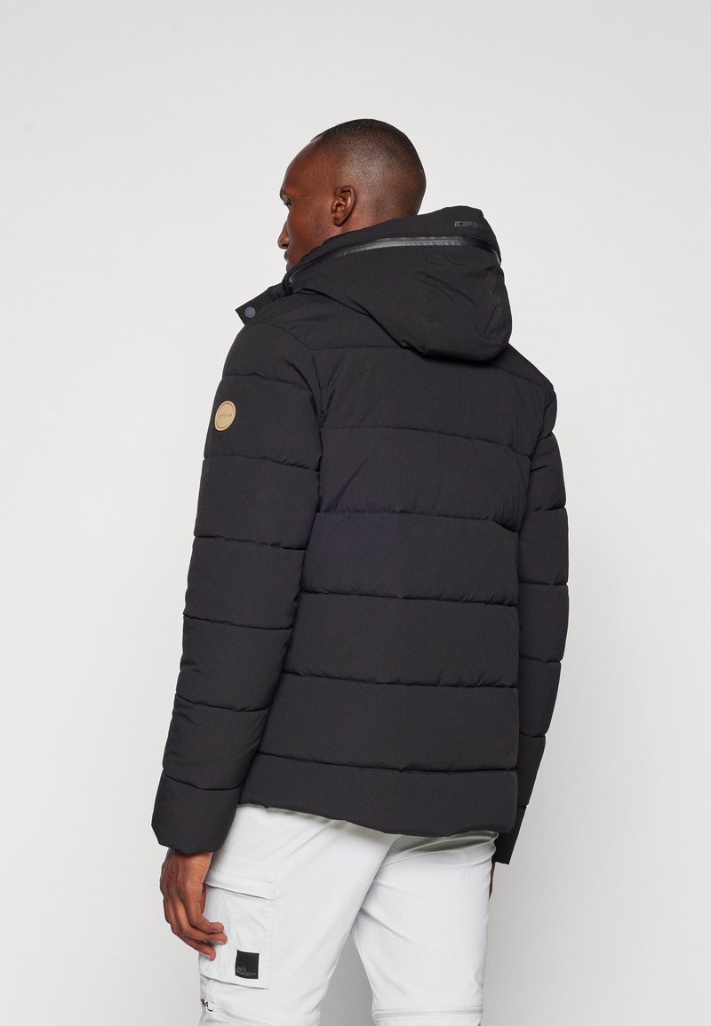 Schwarze Pufferjacke mit Kapuze, ausgestattet mit horizontalen Steppsegmenten und einem Logopatch am Ärmel. Das Material wirkt glatt und robust.