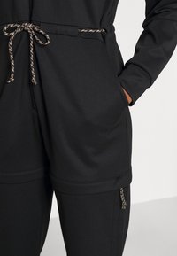 Černý overal s texturovanou tkaninou, přiléhavým střihem, stahovacími šňůrkami v pase se zlatými detaily a bočními kapsami. Rukávy jsou ohrnuté.