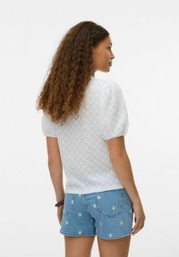 Blouse blanche à manches bouffantes, motif texturé, encolure ronde, associée à un short en denim bleu avec broderie de marguerites.