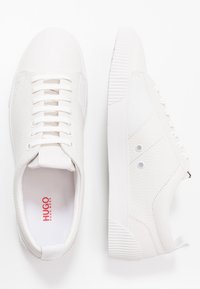 Baskets en cuir blanc avec des panneaux texturés, un design à lacets, des semelles en caoutchouc, et un marquage subtil en rouge sur la semelle intérieure et le côté.