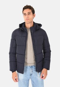 Giacca a vento blu navy con cappuccio, design trapuntato, chiusura con zip e polsini elasticizzati. Indossata sopra un maglione grigio chiaro e abbinata a jeans blu.