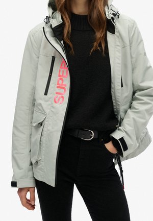 Woman wearing a light grey hooded jacket with pink "SUPER" text, black sweater, black pants, and a black belt with a silver buckle.