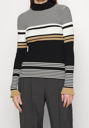 Femme portant un pull côtelé à col haut avec des rayures horizontales noires, blanches et beiges, associé à un pantalon gris foncé.