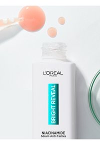 Le sérum Bright Reveal Niacinamide de L'Oréal Paris dans un flacon blanc avec un texte turquoise. Comprend trois gouttes de sérum rose clair à proximité.