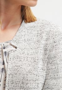 Blazer gris con estampado y tejido texturizado; con solapas de muesca, forma ajustada, combinado con una blusa que presenta un delicado estampado de insectos.