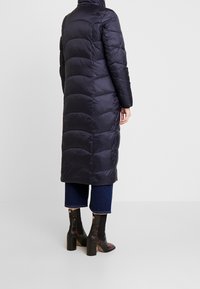 Dlouhý tmavě modrý prošívaný kabát s horizontálním prošíváním, vysokým límcem a dlouhými rukávy. Kombinován s tmavými zvonovými džíny a podpatkovými botami s vzorem.