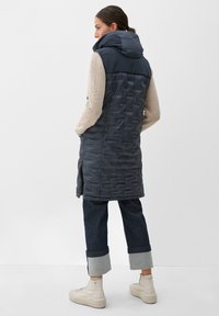 Marineblauer ärmelloser Steppweste mit Kapuze, mit einer strukturierten, gesteppten Optik und seitlichen Reißverschlusstaschen, getragen über einem beigen Strickpullover und Jeans.