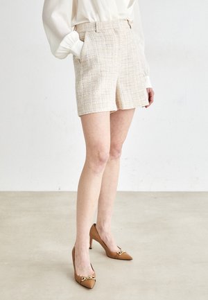 Personne portant un short texturé couleur crème, une blouse blanche à manches longues et des talons hauts pointus marron avec un détail chaîne dorée.