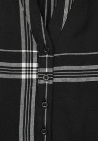 Camicia a quadri bianco e nero, con scollo a V, sei bottoni neri e un motivo di tessuto testurizzato con linee orizzontali e verticali.