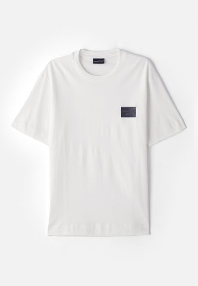 White cotton t-shirt with a round neckline and short sleeves. Features a dark rectangular fabric patch on the left chest area.