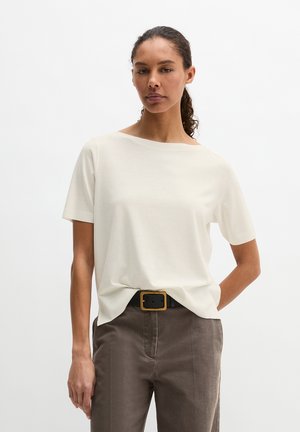 Vrouw met krullend haar, samengebonden, die een effen wit T-shirt met korte mouwen draagt, ingestopt in een hoge taillebruine broek met een zwarte riem.