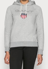 Sudadera gris con capucha y bolsillo canguro, con un logo texturizado de "GANT" y un diseño de escudo en rojo, blanco y azul en el pecho.