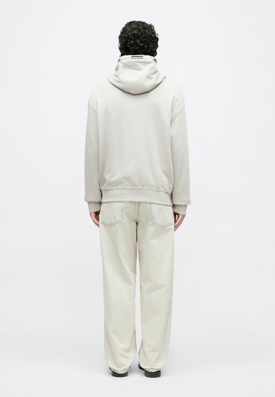 Ανοιχτό γκρι φούτερ με κουκούλα και κορδόνι, συνδυασμένο με φαρδιά, ανοιχτόχρωμα τζιν παντελόνια. Η υφή φαίνεται απαλή και καθημερινή.