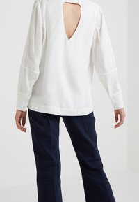 Blusa bianca con maniche lunghe e scollatura a V profonda sul retro, con polsini bottonati e una texture liscia; indossata con pantaloni blu navy.