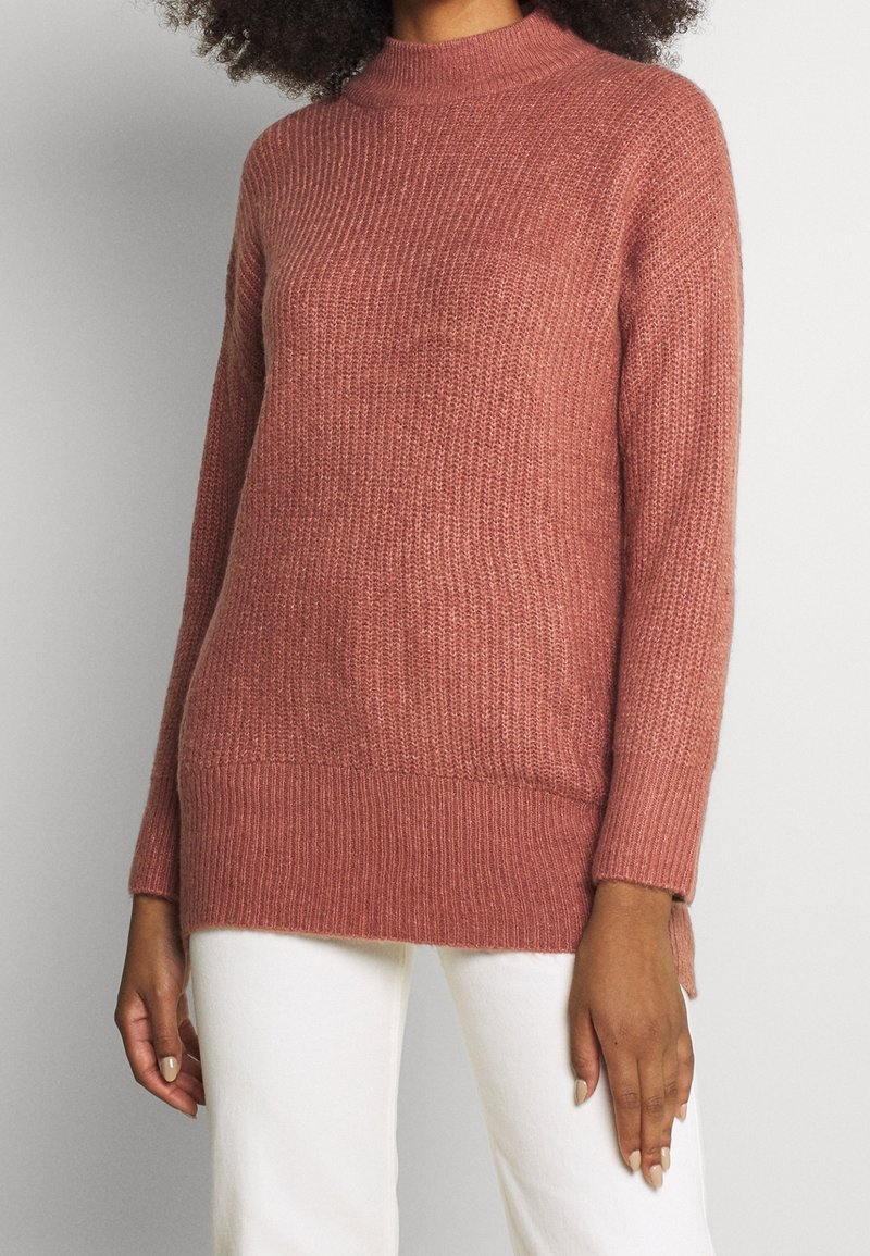 Stickad tröja i dammros med hög krage, ribbad textur och sidofickor. Har långa ärmar och en avslappnad passform.