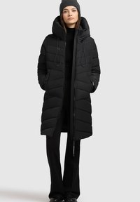 Ung kvinde iført sort pufferjakke med hætte, sort hue, sort turtleneck, bukser og sko, stående foran en grå baggrund.