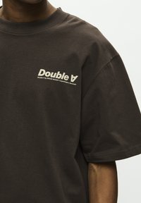 Camiseta de color marrón oscuro con mangas cortas y logo beige "Double A by WOOD WOOD Copenhague, Dinamarca" en la parte superior izquierda del pecho.