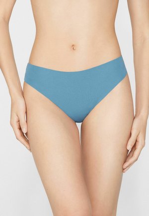 Femme portant un sous-vêtement bleu seamless taille basse, visible du milieu du torse jusqu'en haut des cuisses sur un fond uni.