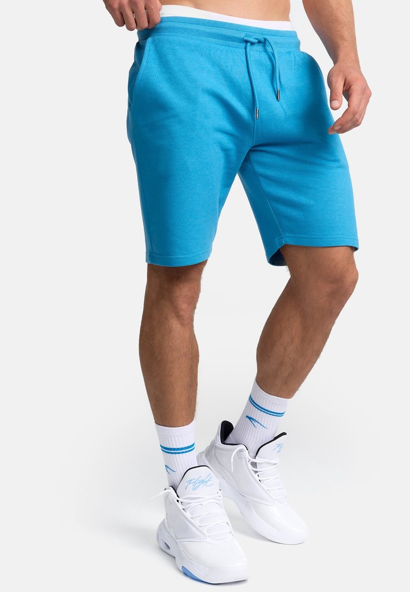 Shorts turquoise en mélange de coton avec une taille élastique et un cordon de serrage. Comprend des poches latérales et une coupe décontractée. Associés à des baskets blanches.
